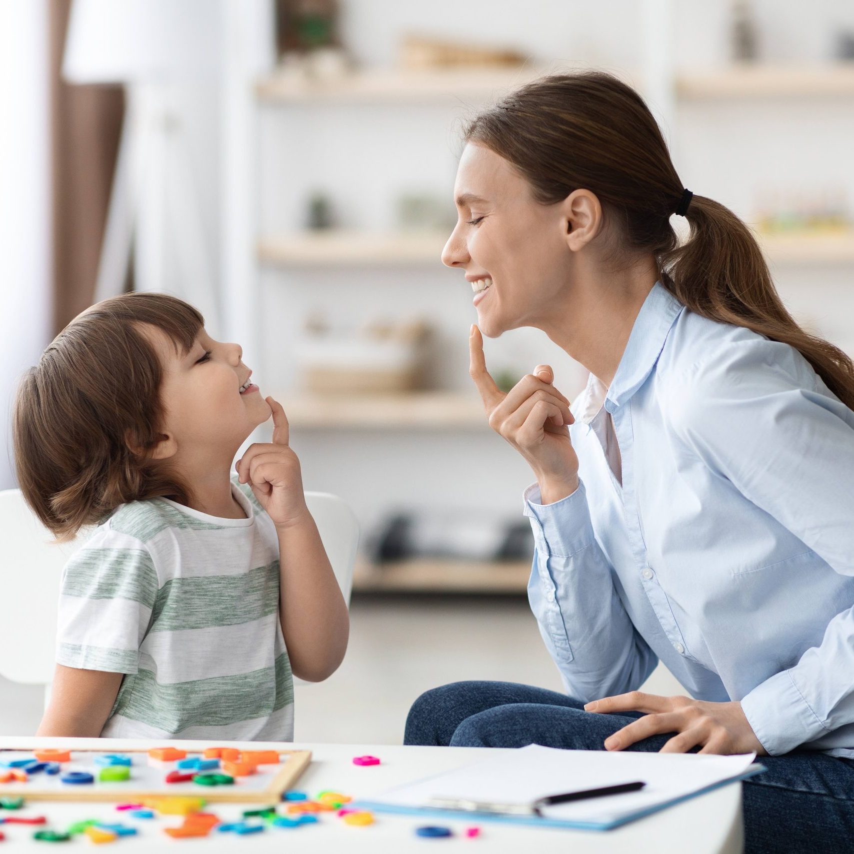 Speech training for kids. Professional woman specialist training with little boy at cabinet, teaching him right articulation exercises, side view